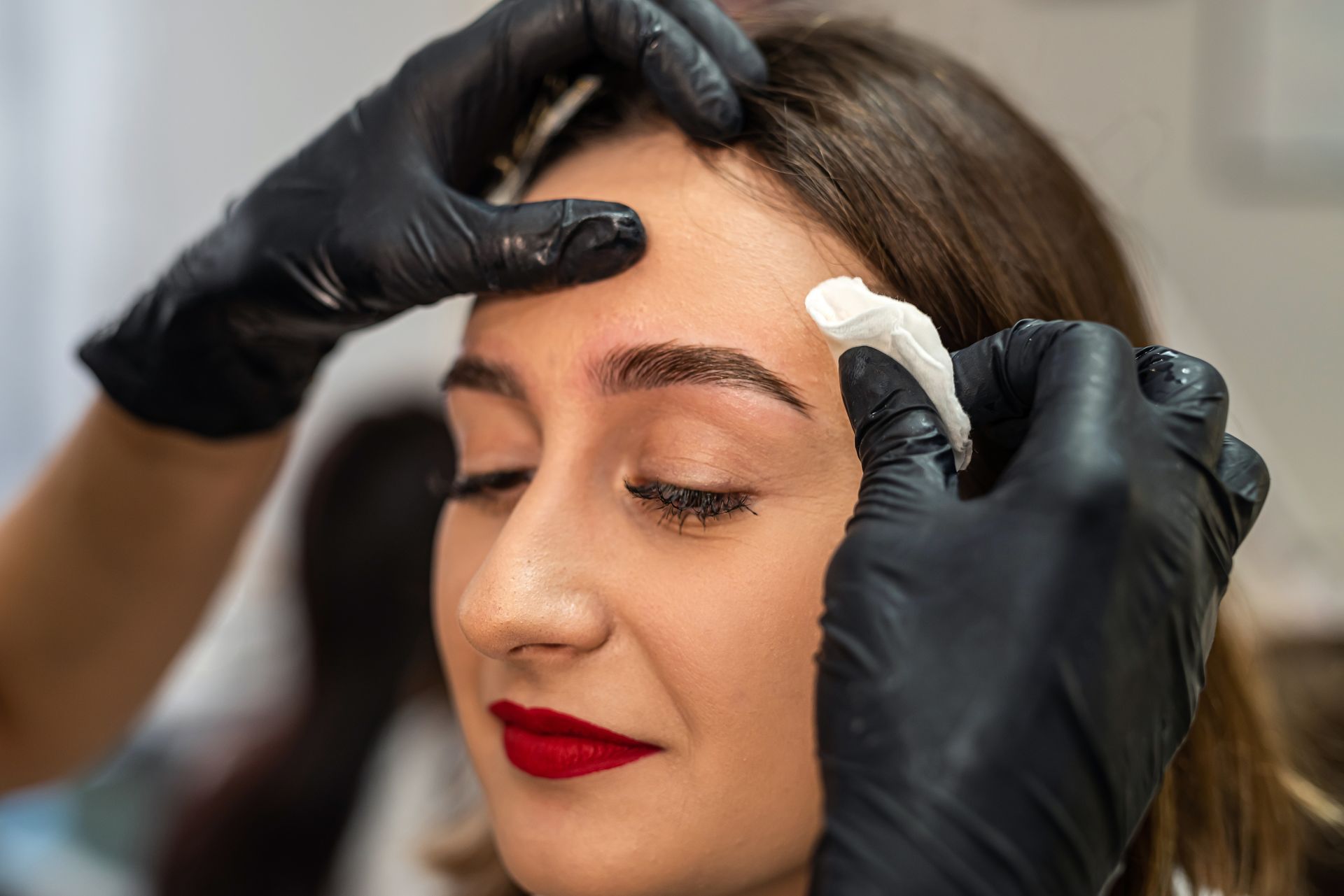 Woman having her eyebrow waxed at a salon; red lipstick, black gloves, close-up.