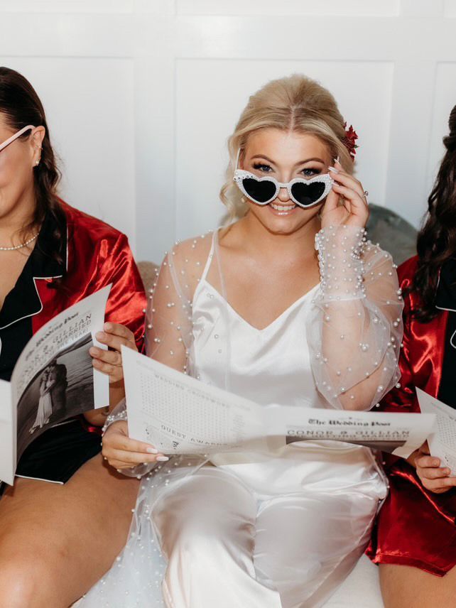 Bride wearing heart-shaped sunglasses, holding a newspaper, with bridesmaids in red pajamas.