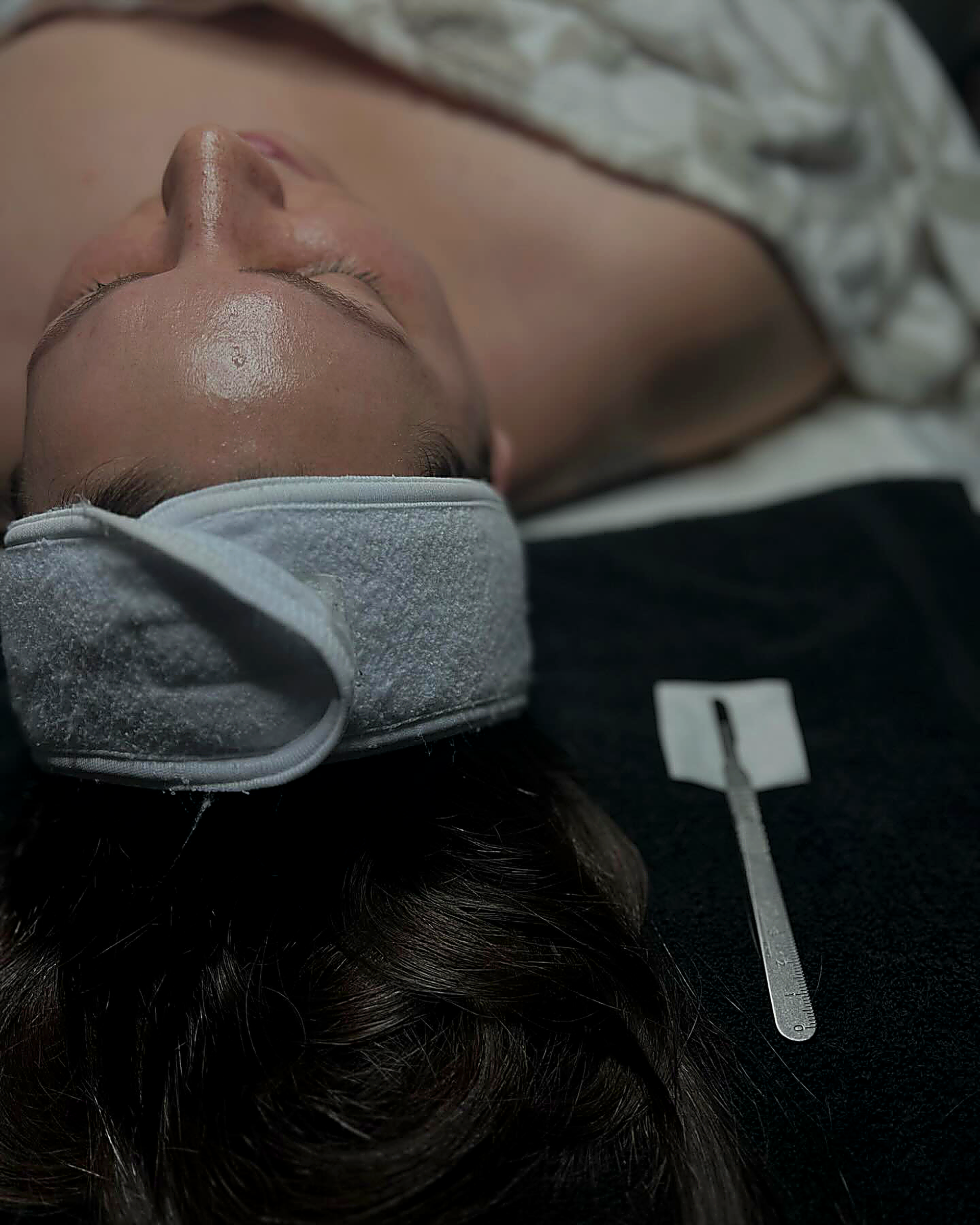 A person lying down with a white headband, receiving a facial treatment with a scalpel on a black surface.