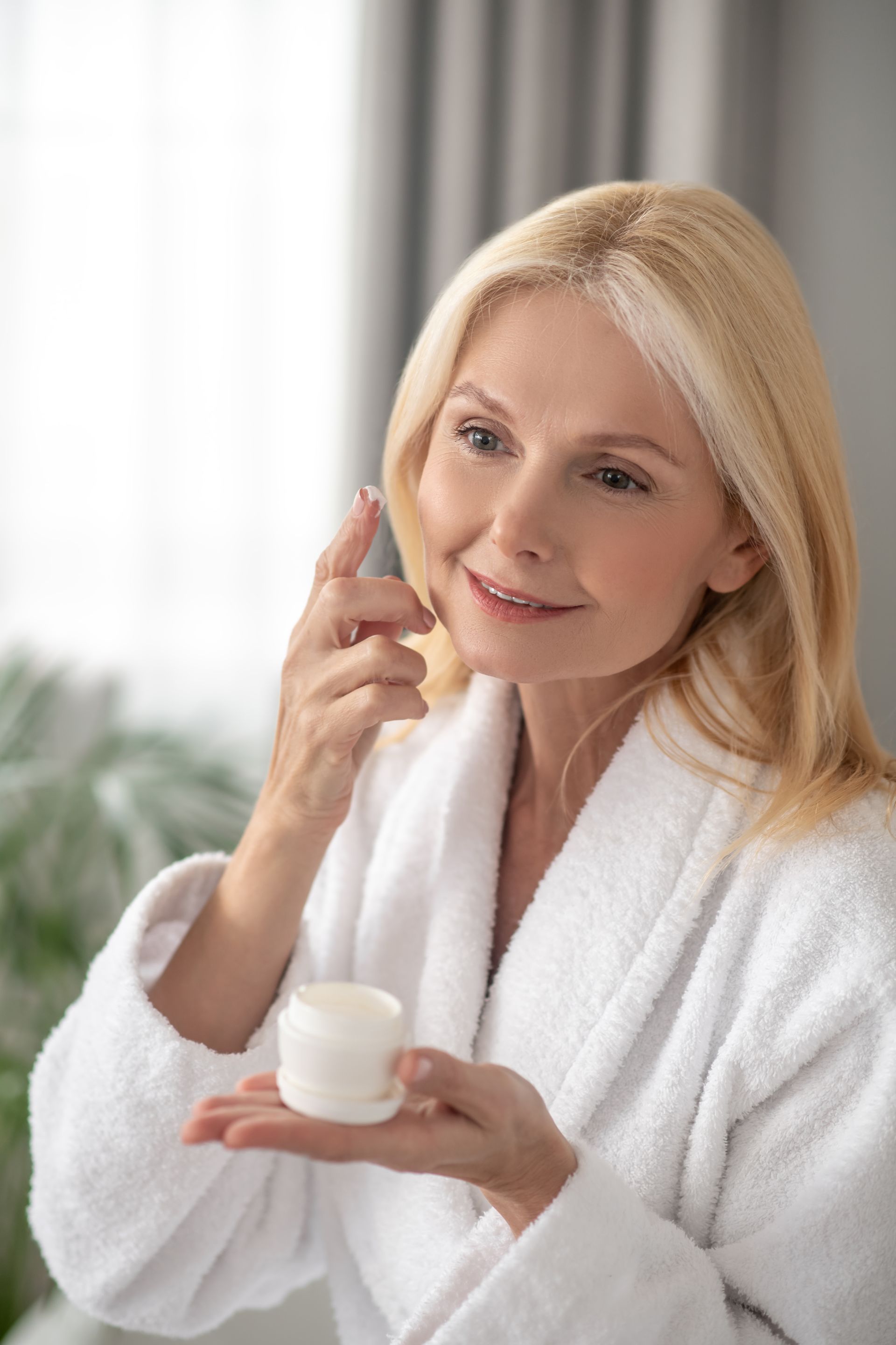 A woman in a bathrobe is applying cream to her face.