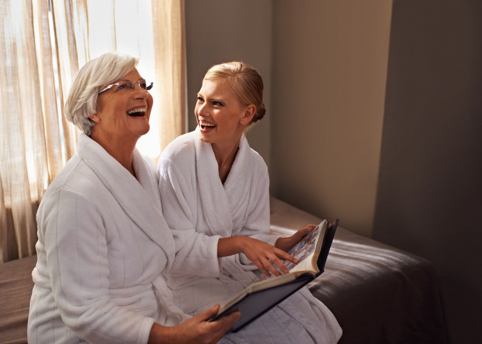 Two people in white robes sit on a bed together, laughing while looking at an open photo album.
