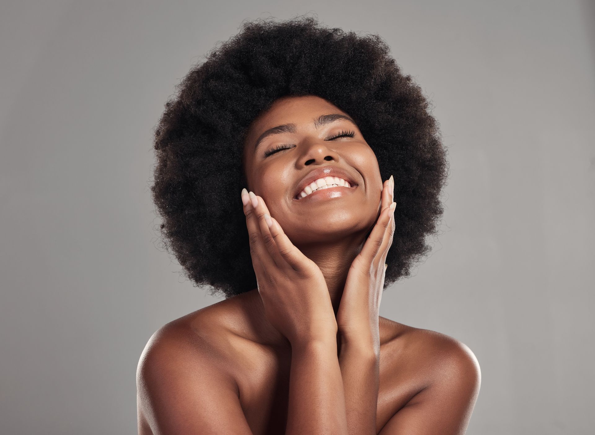 A woman with an afro is smiling and touching her face.