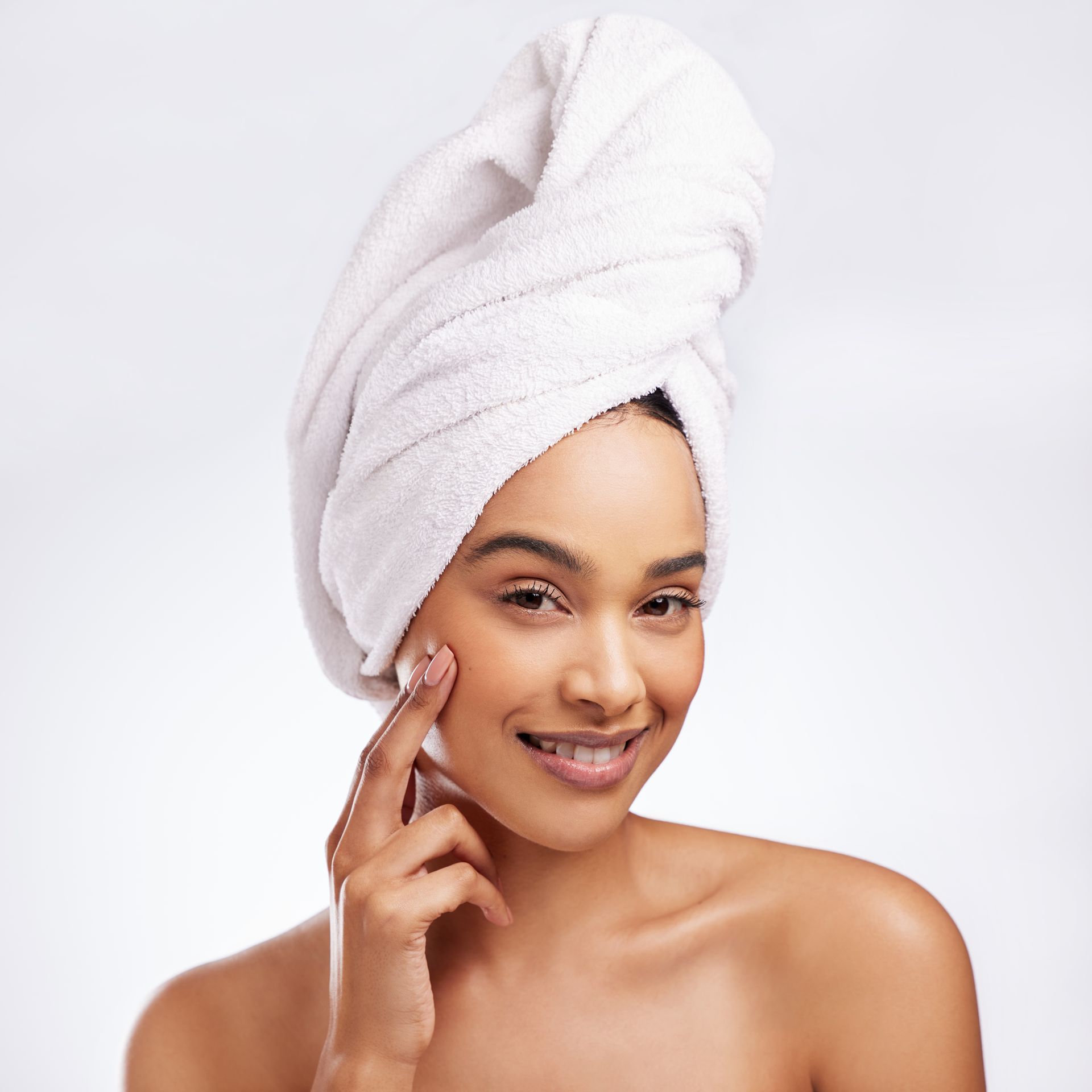 A woman with a towel wrapped around her head is smiling and touching her face.