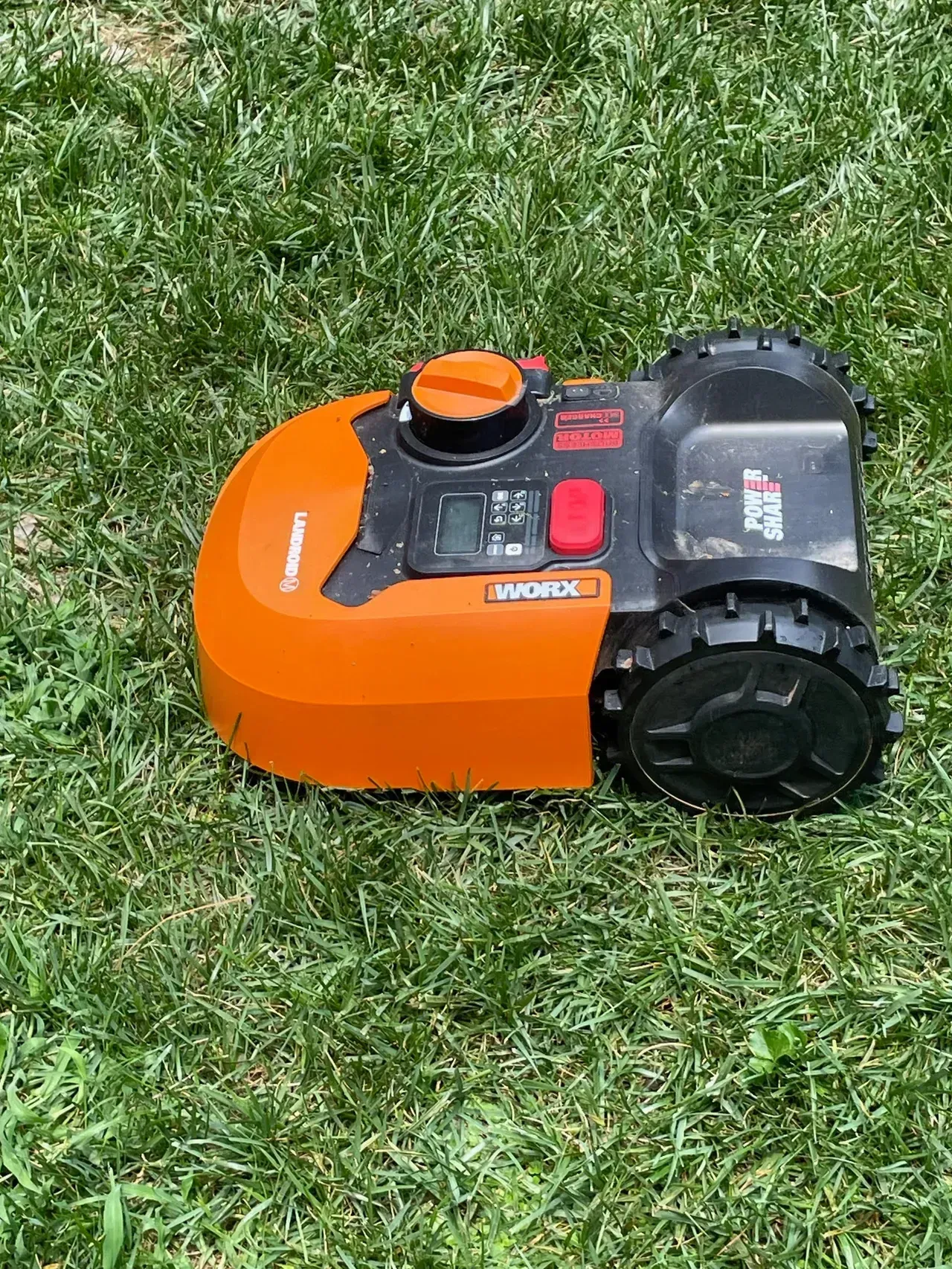 A dark grey, futuristic robotic lawnmower sits on vibrant green grass against a plain, minimalist white wall.