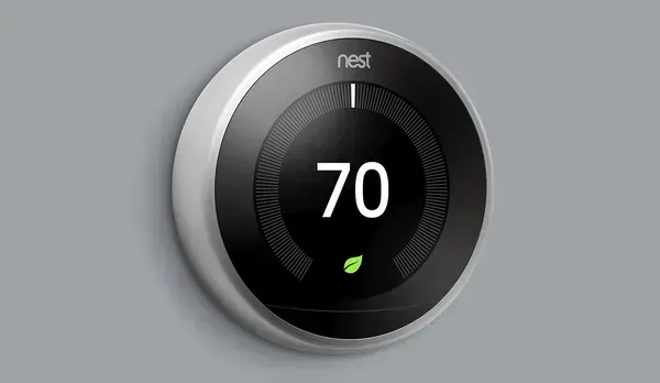 A person's hand adjusts a white, circular smart thermostat mounted on a plain, light-colored wall.