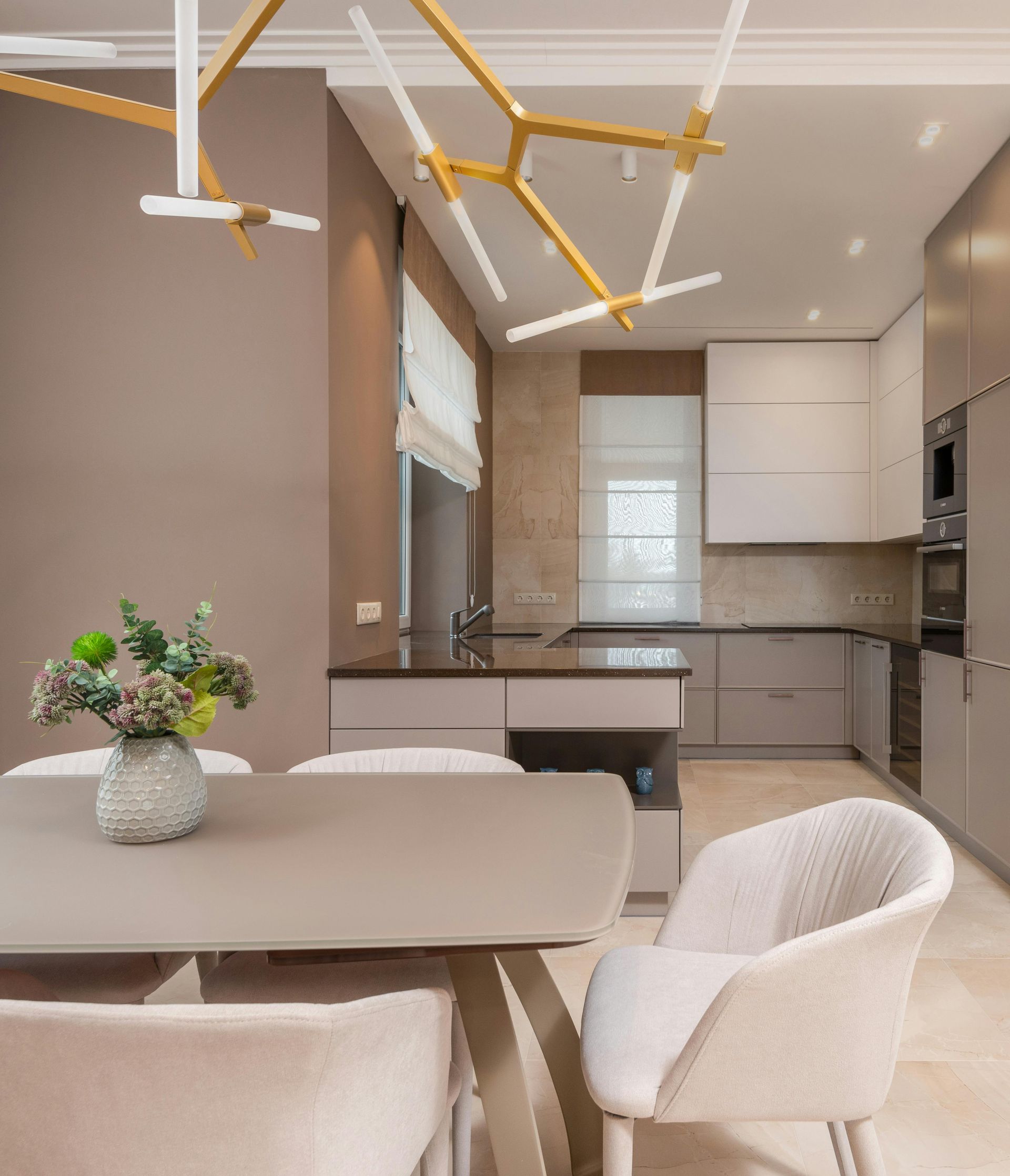 Modern kitchen and dining area with neutral tones. Dining table with chairs, overhead light fixture, and kitchen in background.