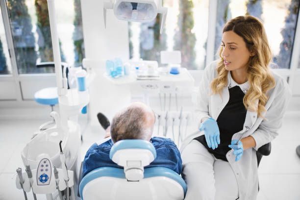 Dentist in white coat and gloves talking to a patient in a dental chair.