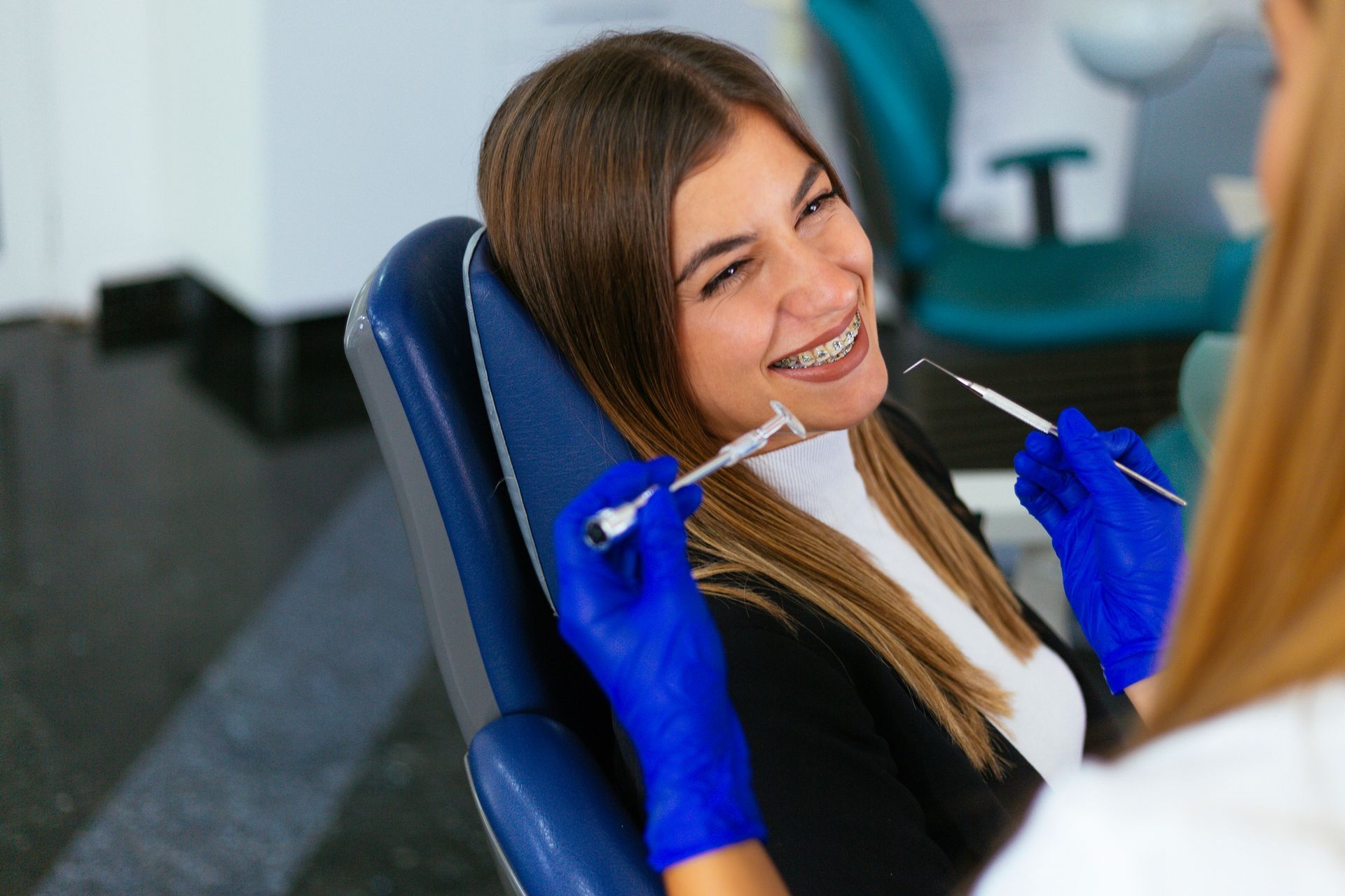 Woman with braces smiles during a dental checkup. Dentist holds instruments, examining teeth.
