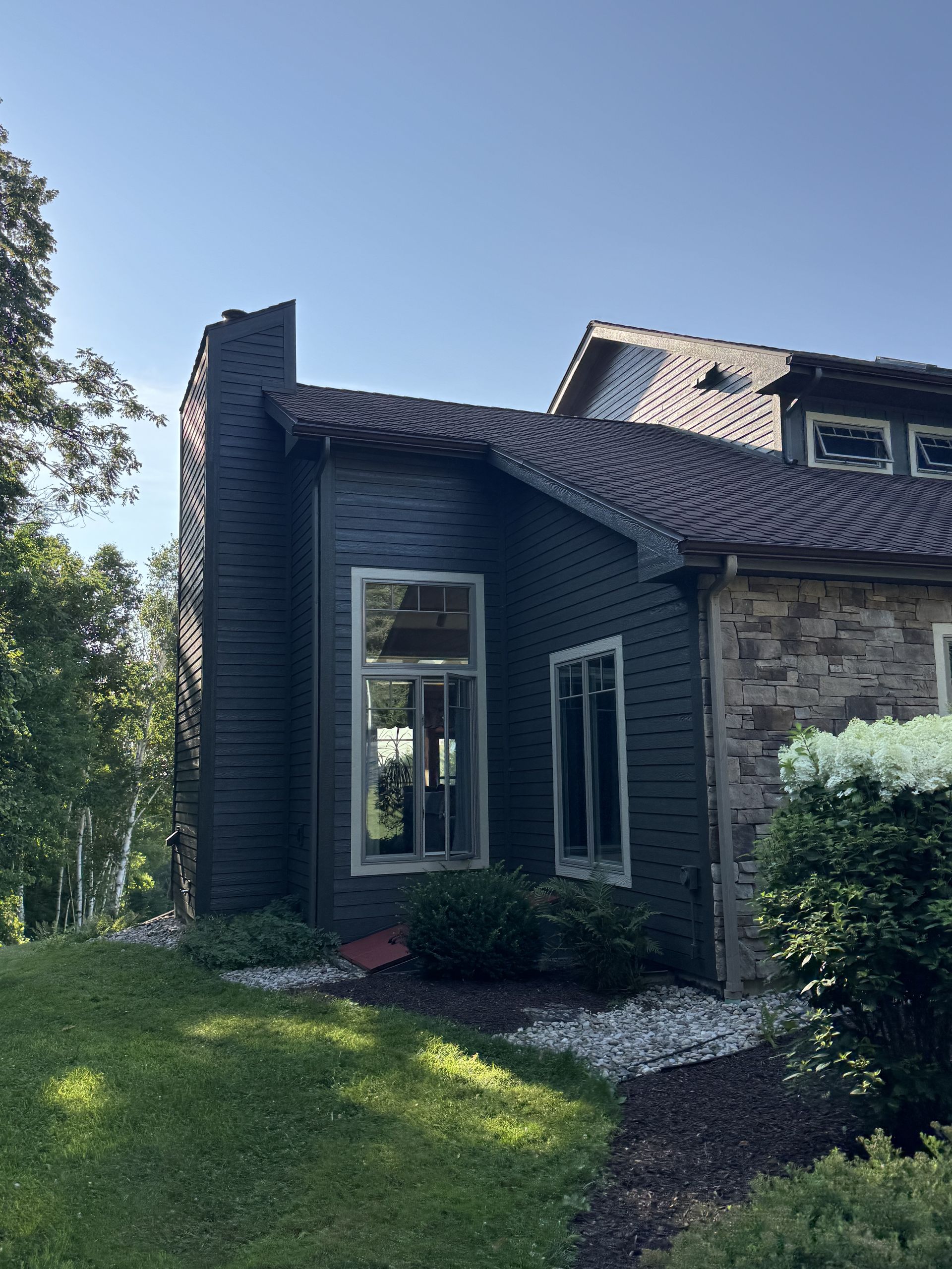 Dark-colored house with a tall chimney. Green grass in front, blue sky in the background.