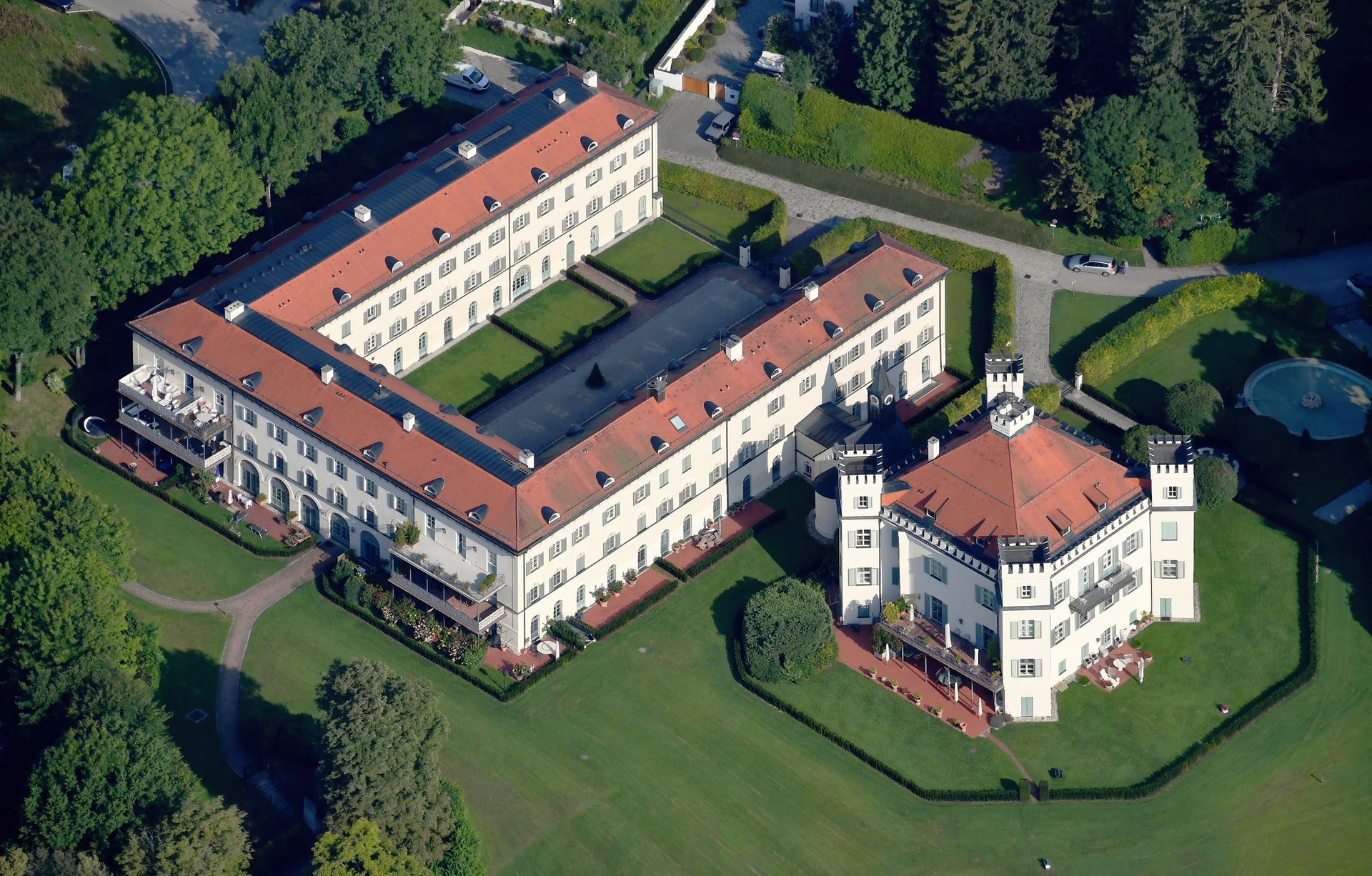 Luftaufnahme eines weißen Gebäudekomplexes mit roten Dächern und einem kleinen Innenhof, umgeben von Bäumen und Gras.