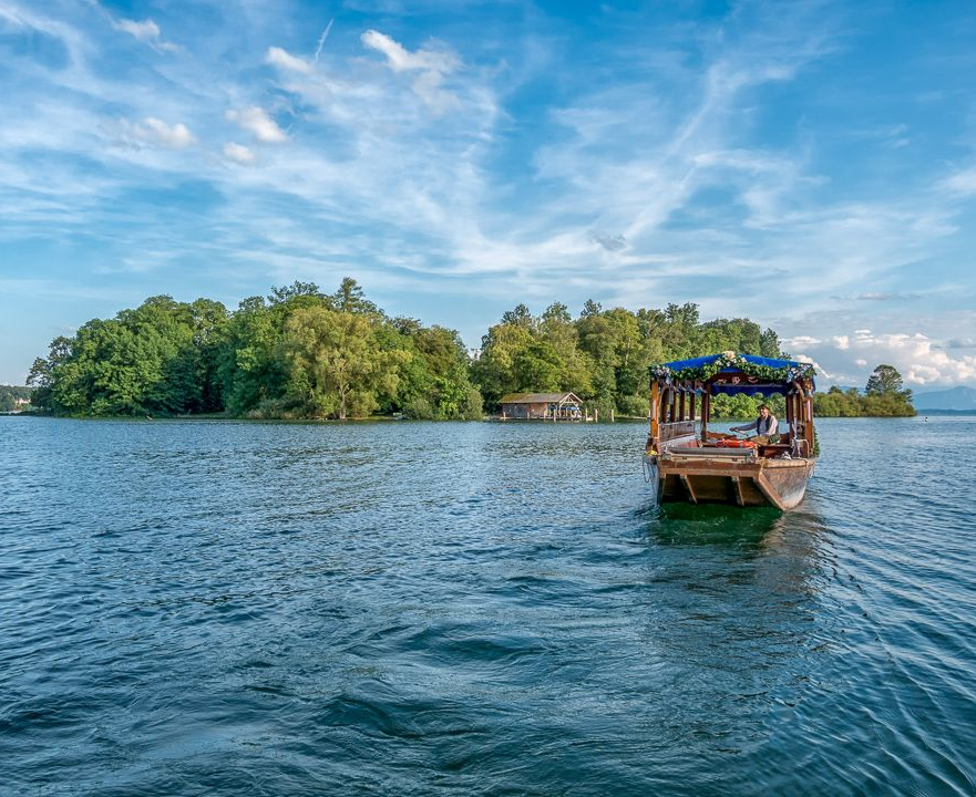 Schattiger Park am Seeufer mit einer Holzbank, einem Segelboot auf ruhigem Wasser und Bäumen, die den Blick einrahmen