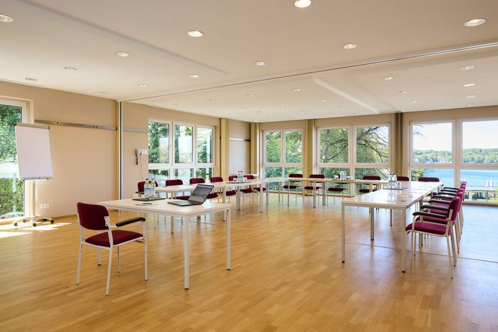 Konferenzraum mit großen Fenstern mit Blick auf einen See und ein Gebäude; rote Stühle, beige Vorhänge und weißer Tisch.