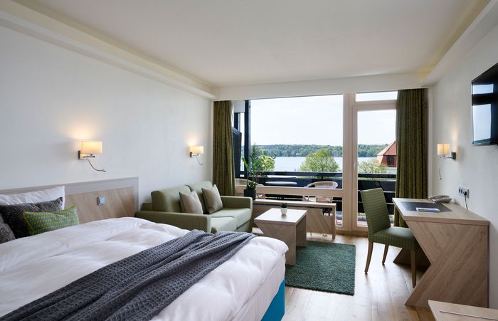 Hotelzimmer mit Bett, Balkon und Seeblick. Blaue Vorhänge, Holzvertäfelung, Fernseher an der Wand.