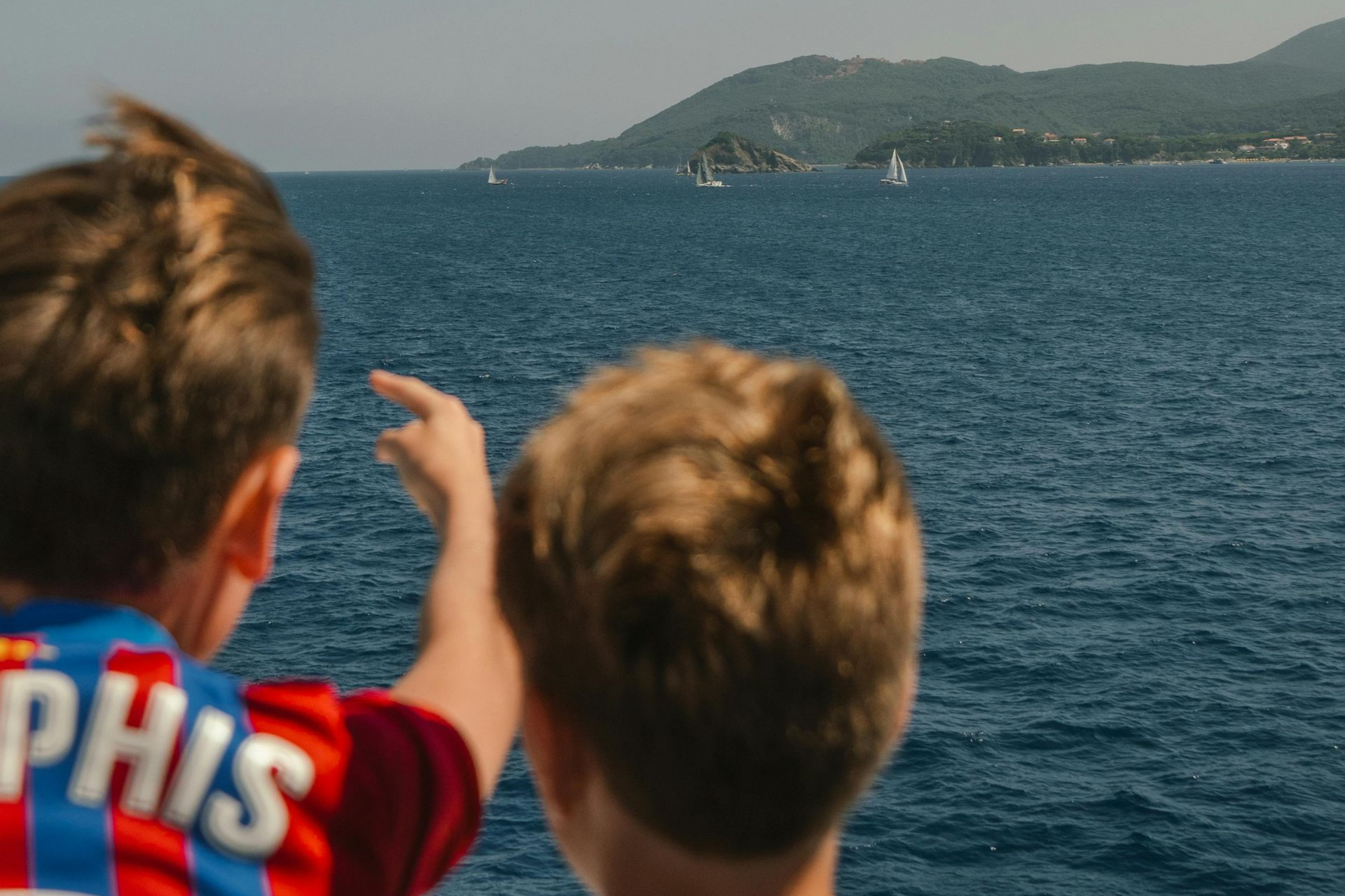 Zwei Jungen schauen aufs Meer hinaus, einer zeigt auf Segelboote in der Ferne.