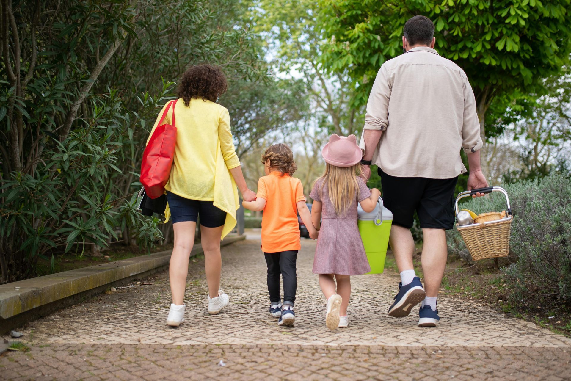 Eine Familie spaziert Händchen haltend und mit einem Picknickkorb in der Hand auf einem Weg in einem Park.