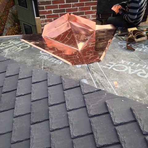 A man is sitting on top of a roof with a copper chimney.