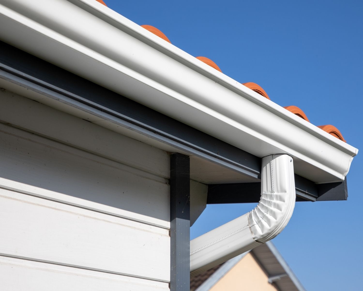 White gutters and downspout on a house with white siding and orange roof tiles, against a blue sky.