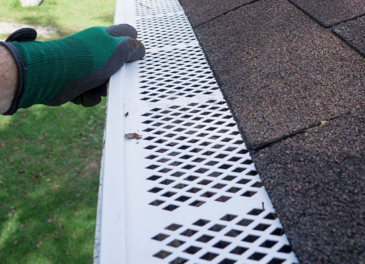 Gloved hand holding a white gutter guard installed on a dark shingle roof, over green grass.