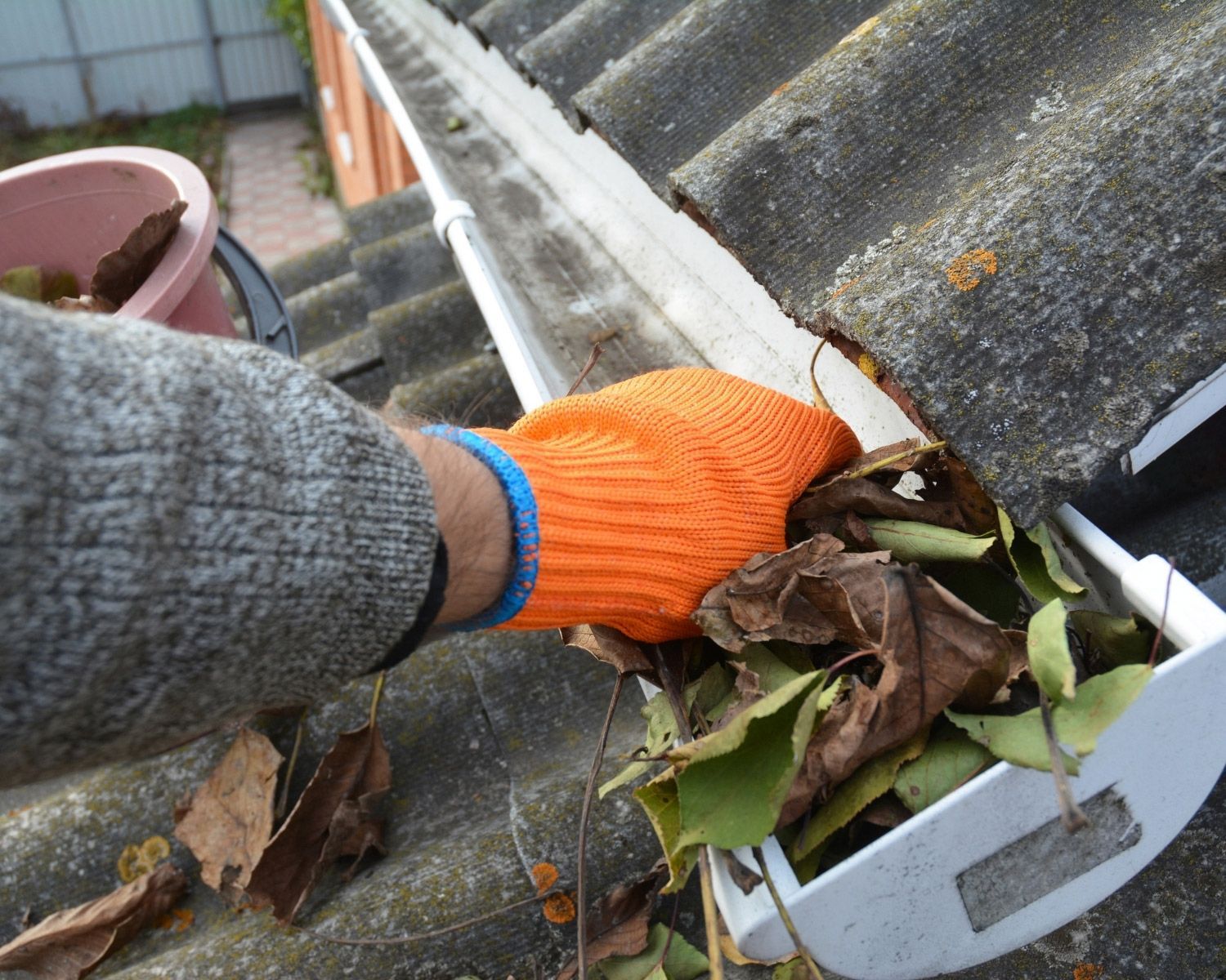 Person in orange glove cleaning leaves from a gutter on a roof.