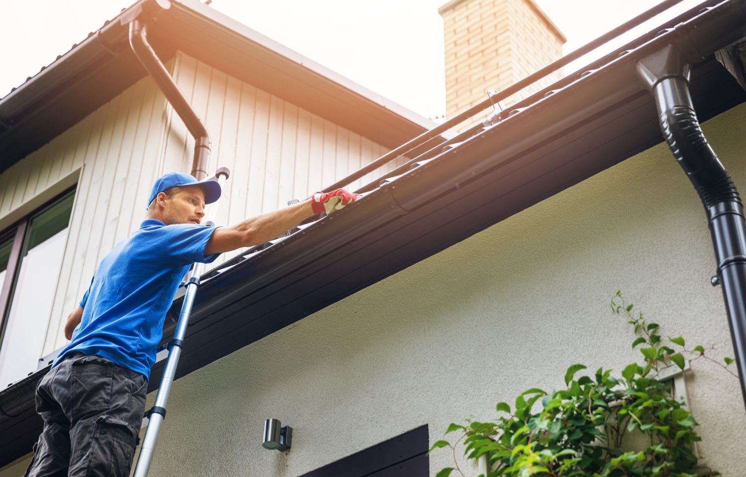 Man on a ladder cleaning a black gutter on a white house.