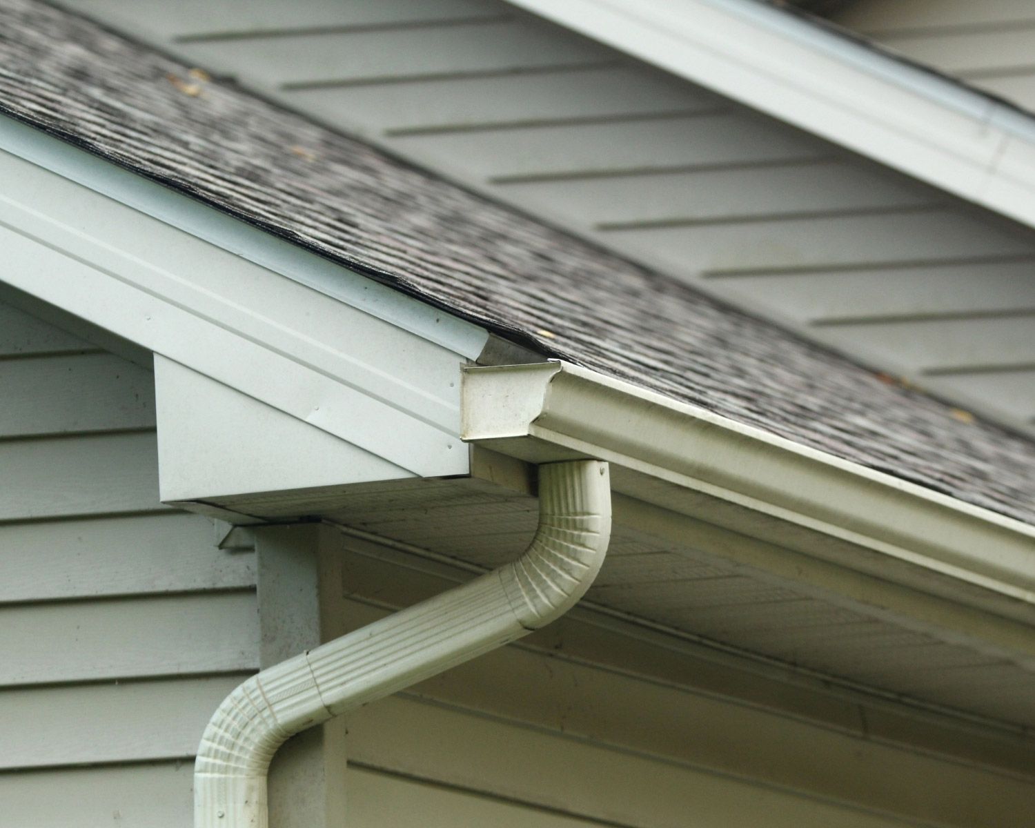 Beige gutters and downspout on a house with gray siding and roof.