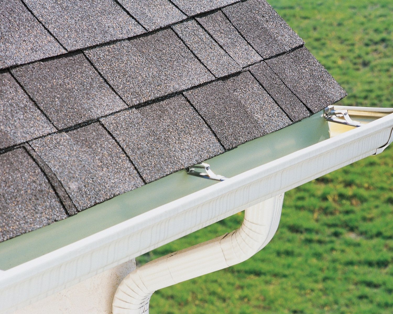 White gutter along a shingled roof, with green grass in the background.