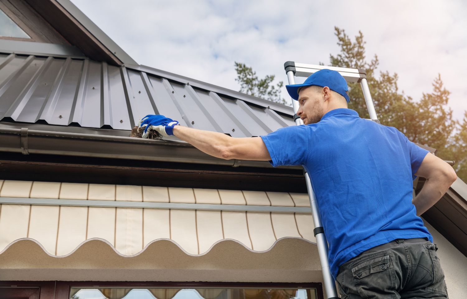 Man on ladder cleaning a gutter on a brown metal roof.