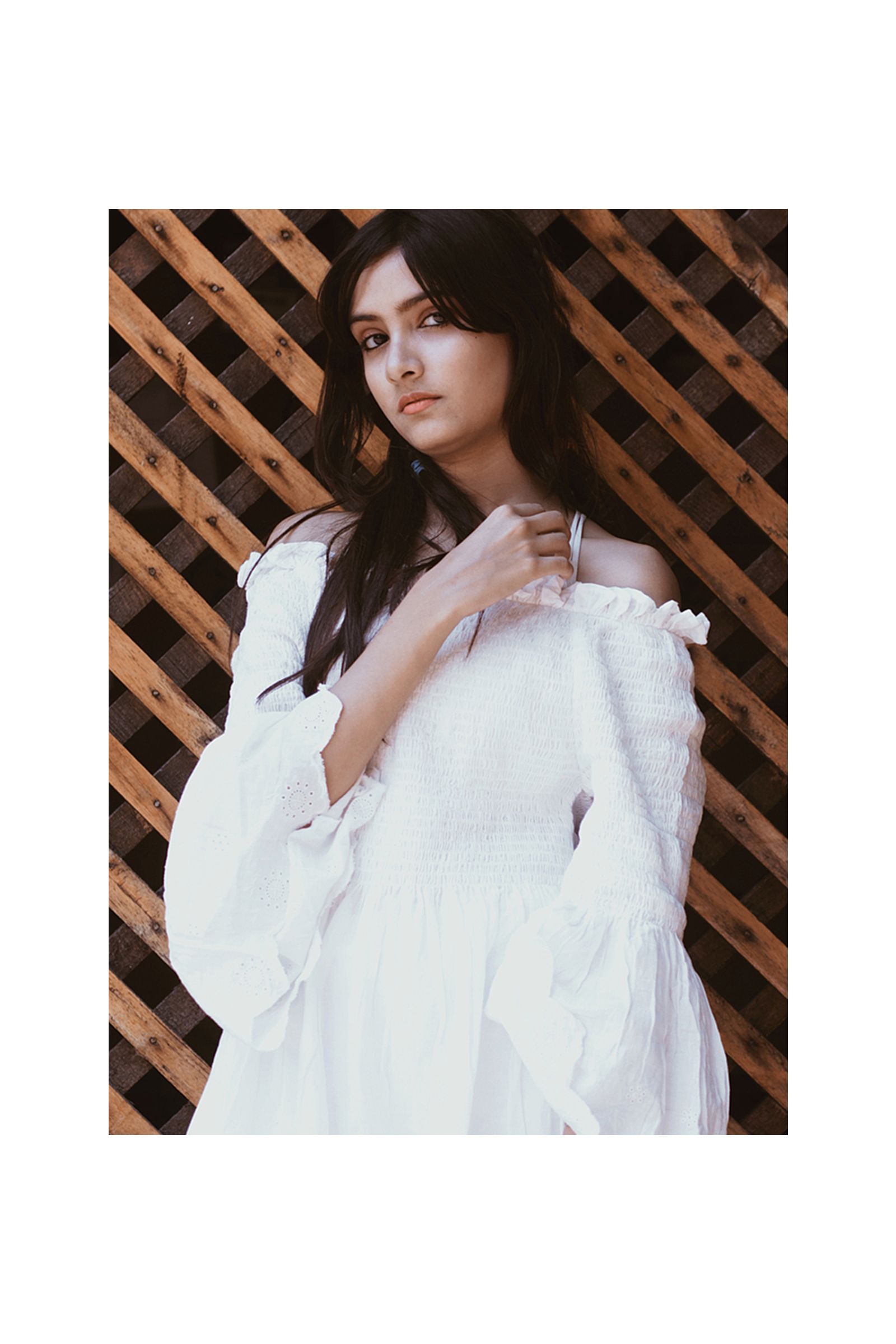 A woman in a white off the shoulder dress is standing in front of a wooden fence.