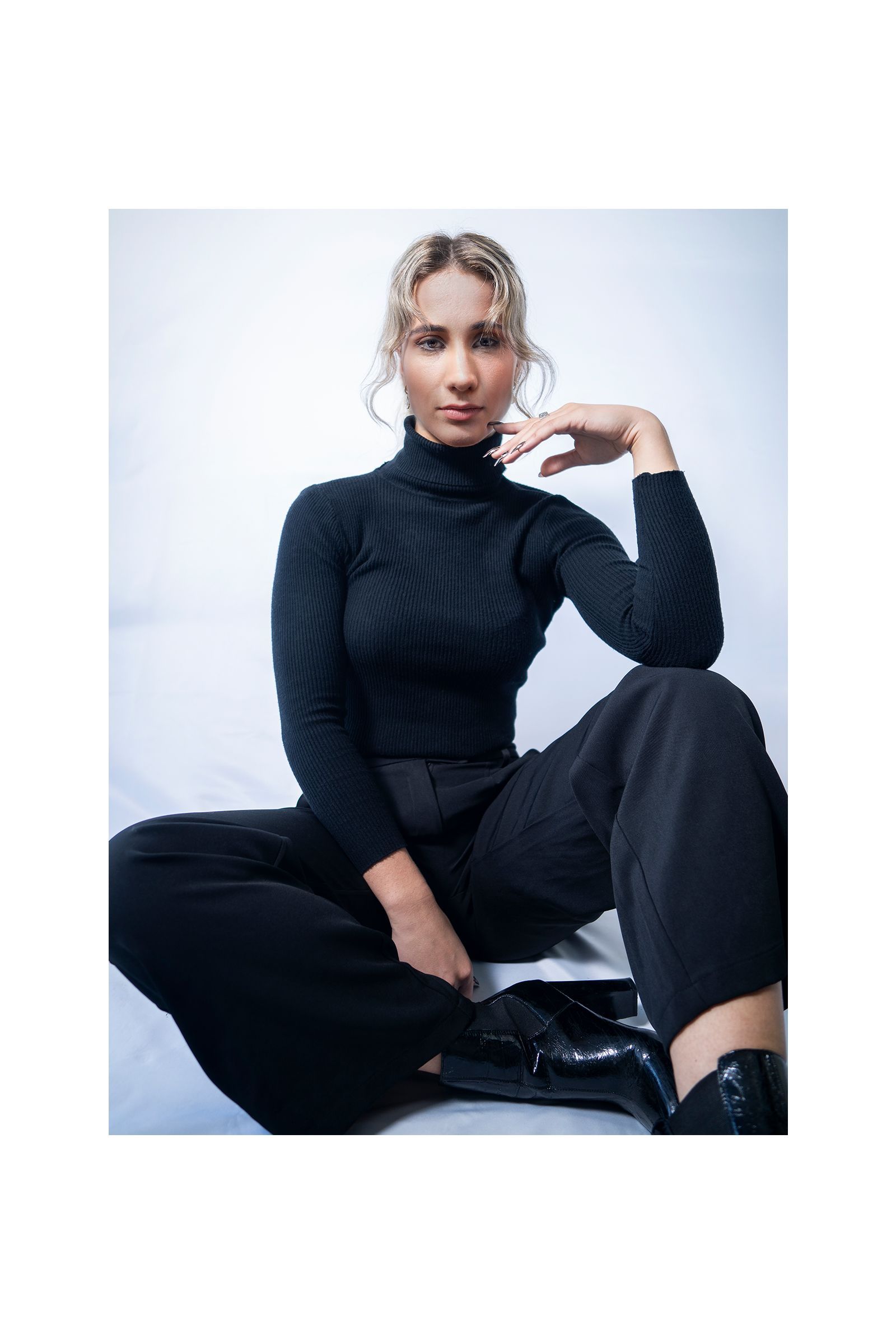 A woman is sitting on the floor wearing a black turtleneck and wide leg pants.