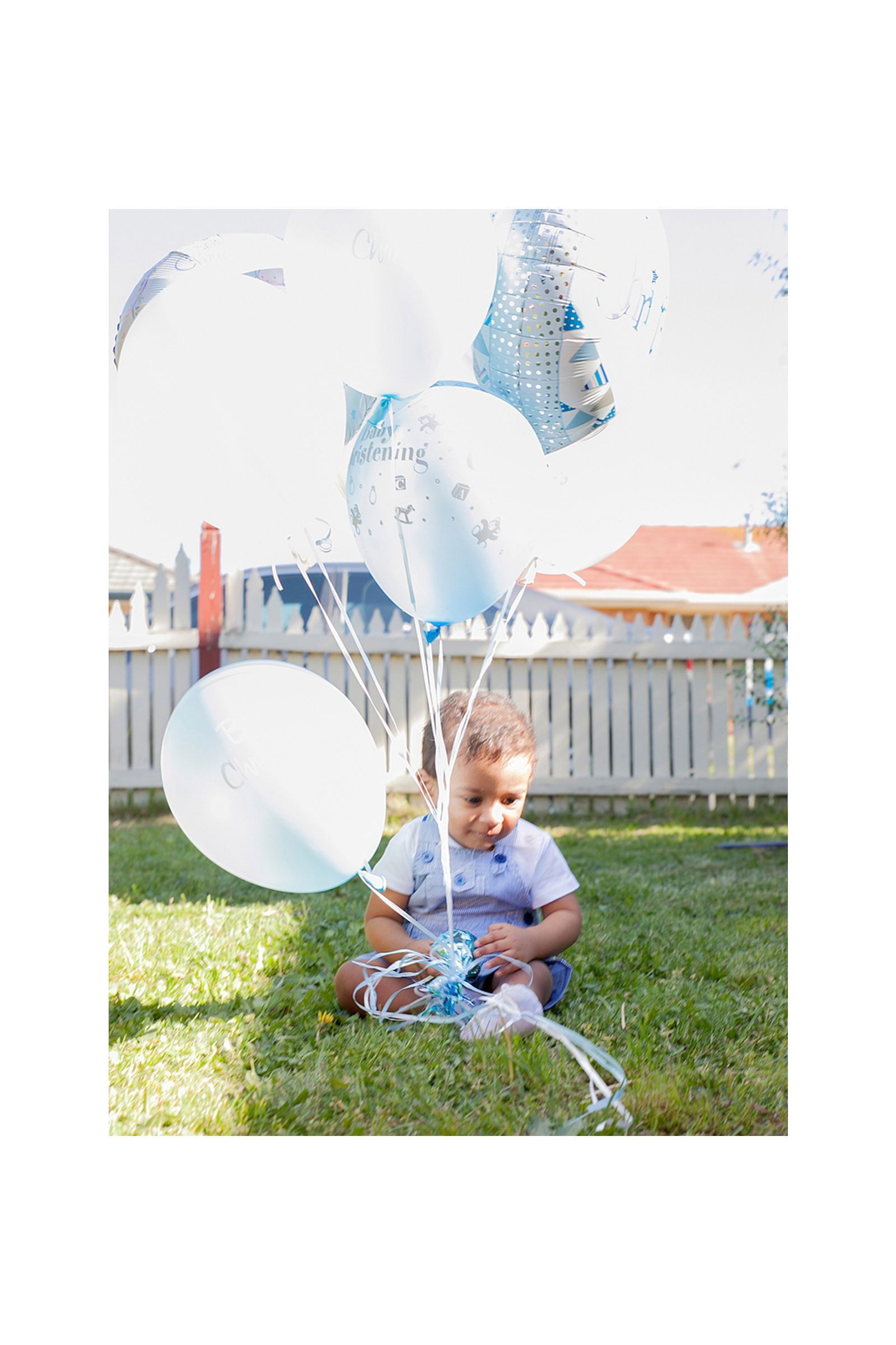 A baby is sitting in the grass holding balloons.