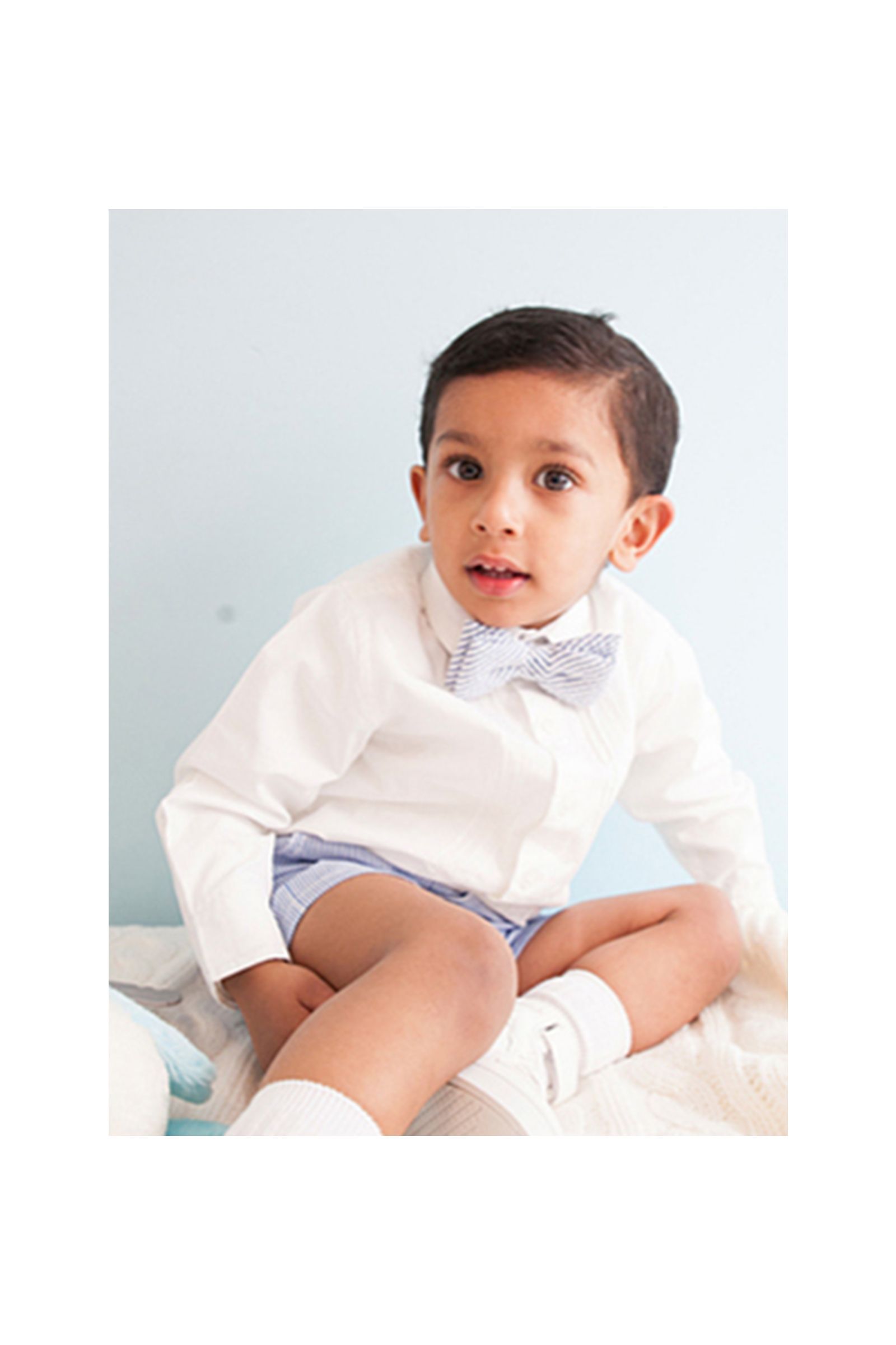 A little boy wearing a white shirt and a blue bow tie is sitting on a bed.