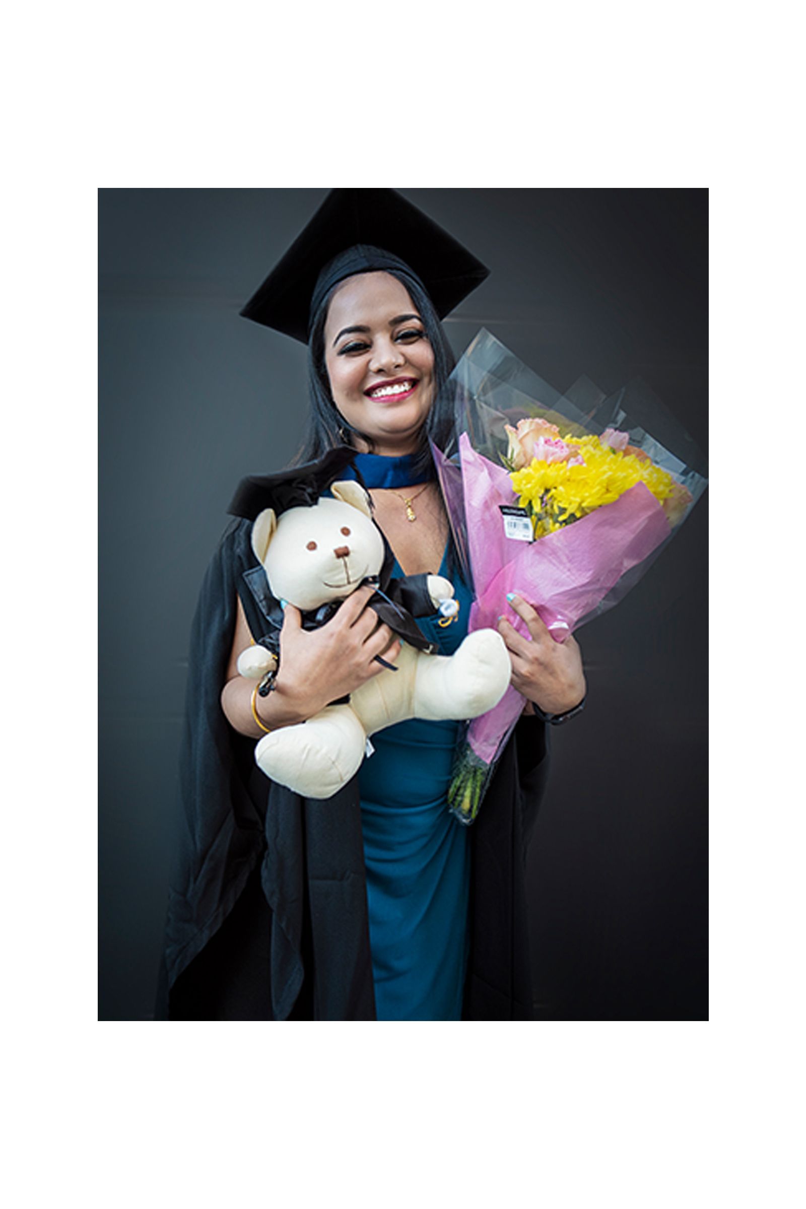 A woman in a graduation cap and gown is holding a teddy bear and a bouquet of flowers.