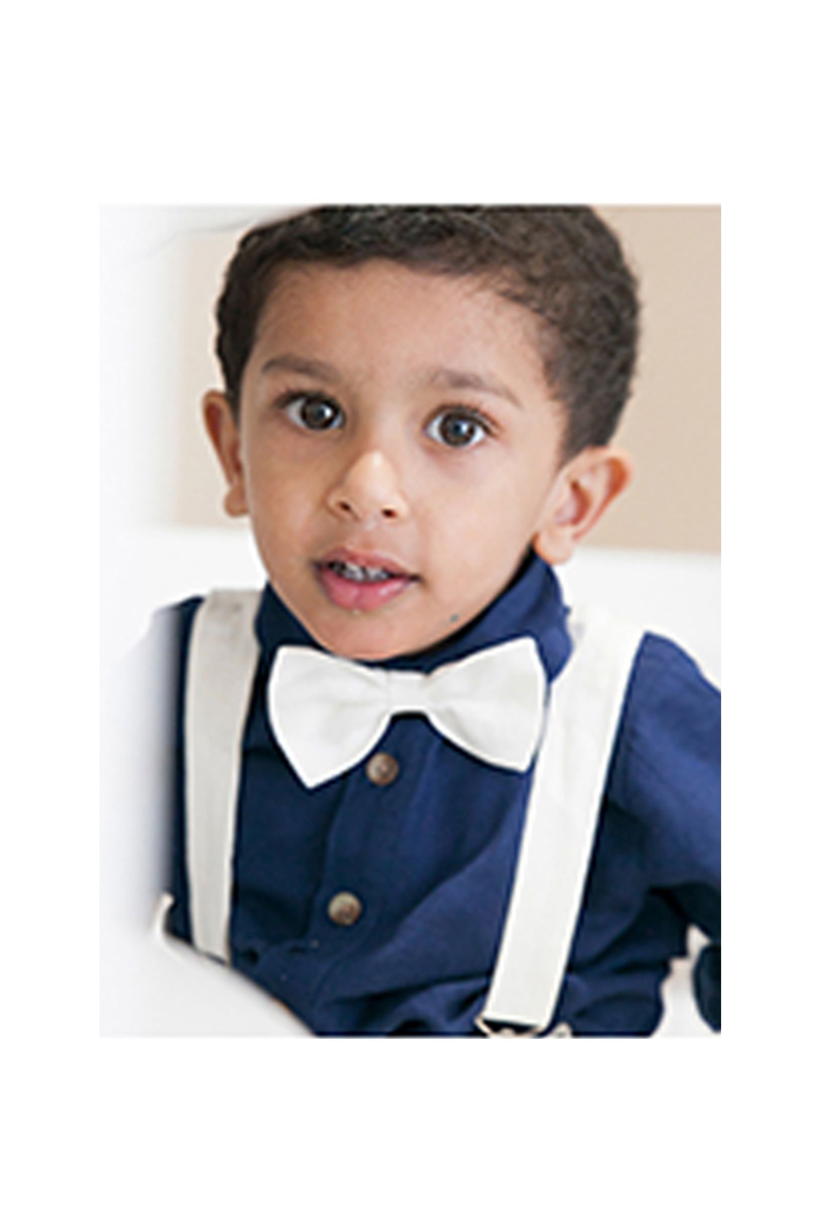 A young boy wearing a bow tie and suspenders is sitting on a couch.