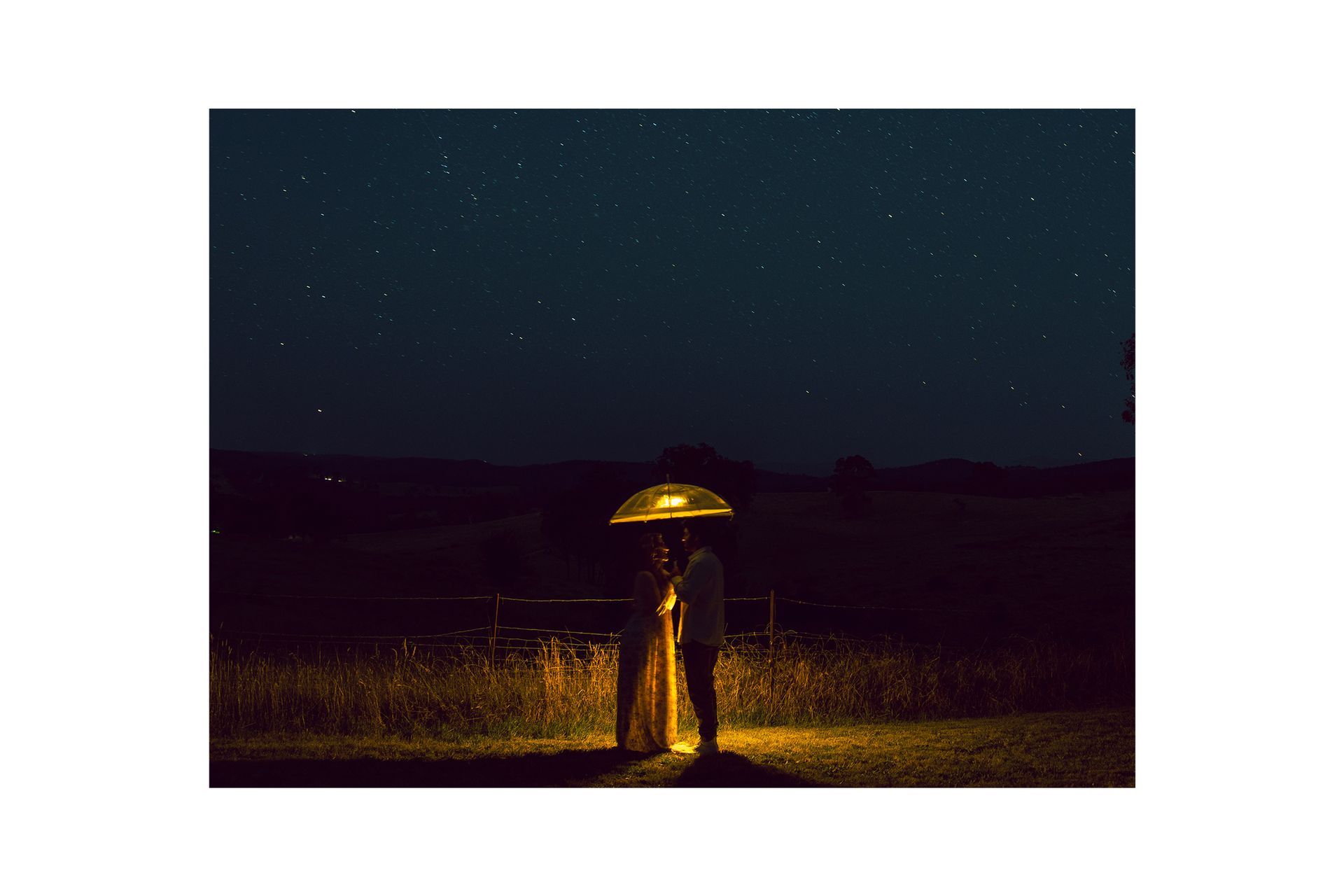 A bride and groom are standing under an umbrella in a field at night.