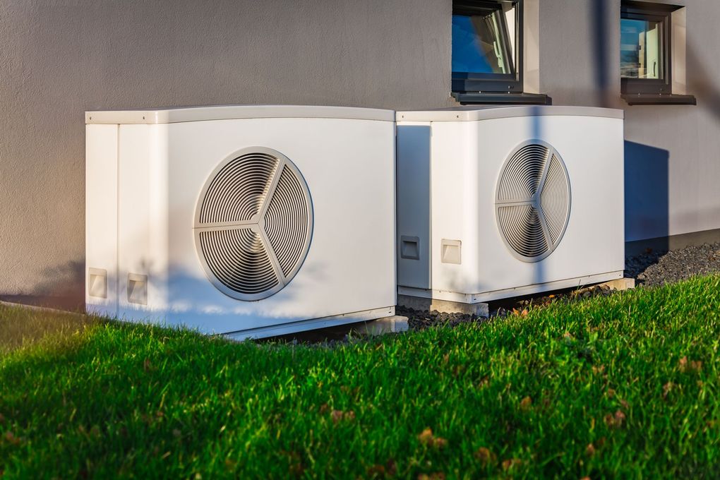 Two white outdoor HVAC units beside a building, with grass in the foreground.
