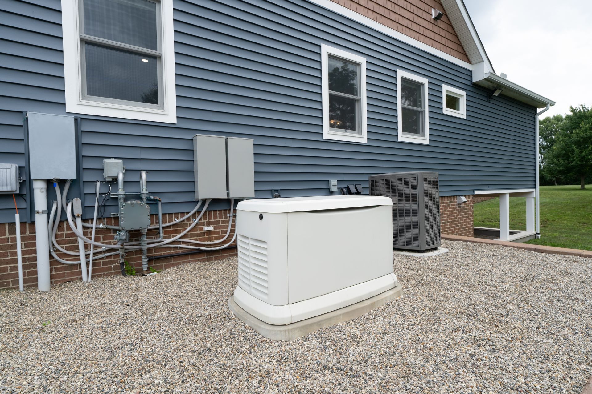 White generator on gravel beside blue house with utility meters and pipes along the wall