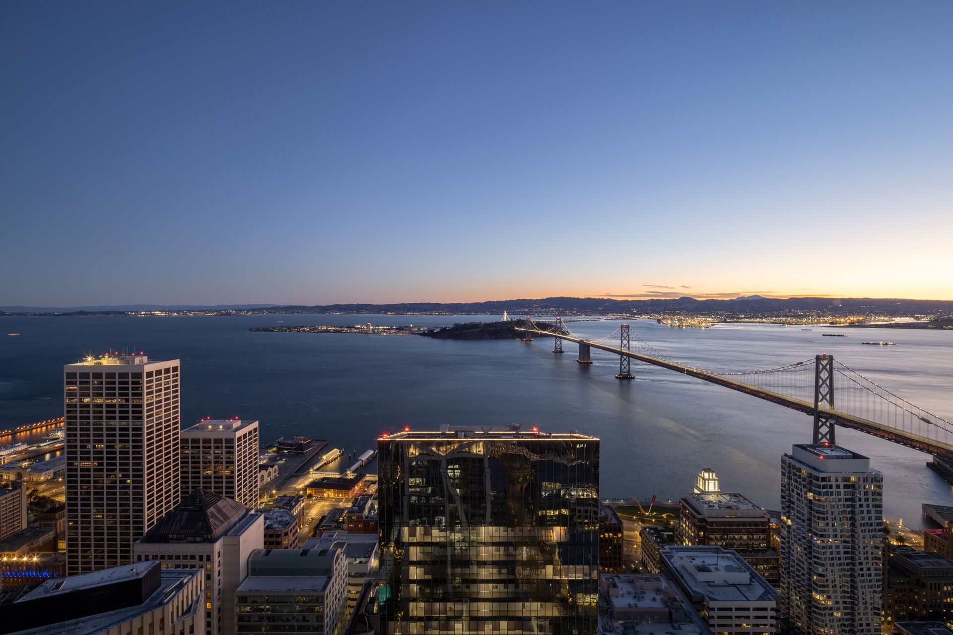 City skyline at dusk with Bay Bridge and water, soft blue sky, building lights reflecting.