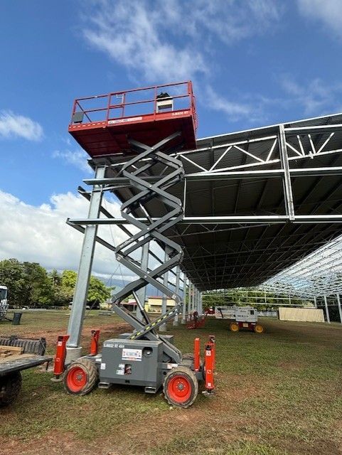 A red scissor lift is extended to reach the metal roof frame of a construction site under a partly cloudy sky.
