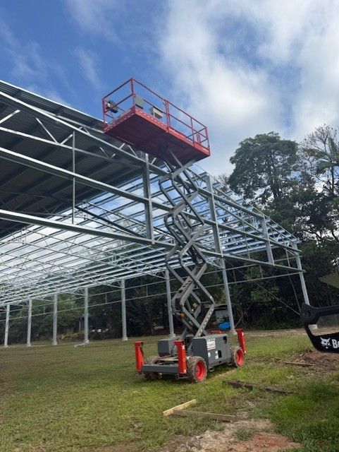 A red scissor lift is extended high to reach the roof framework of an outdoor steel building construction site.