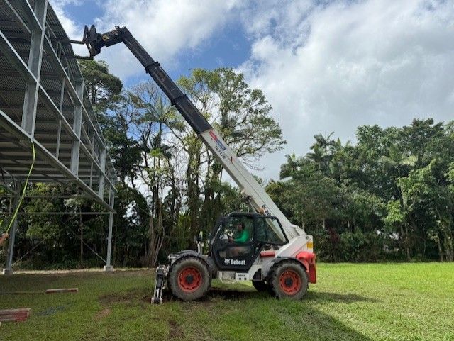 A Bobcat telehandler extends its boom to reach the steel framework of a structure in a grassy field.