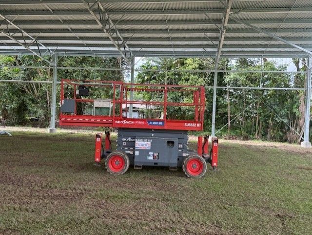A red Snorkel scissor lift stands on a grassy field under a large, open-sided metal structure.