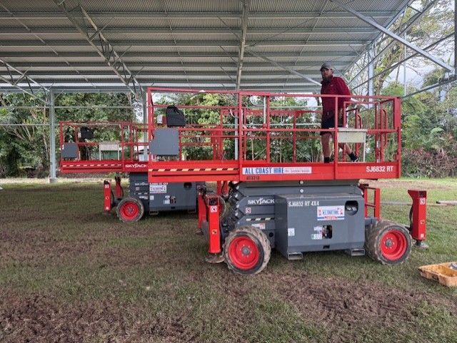 Two red and gray scissor lifts parked under a metal-roofed shelter on a grassy field, with a person standing on one.