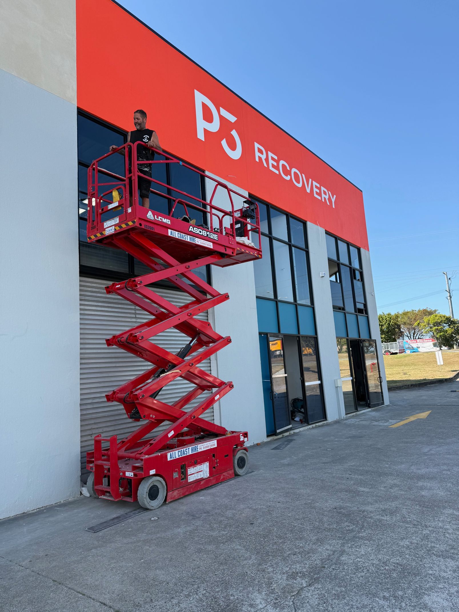 A worker stands on a red scissor lift, performing maintenance on the exterior of a building with a large red P3 RECOVER sign.