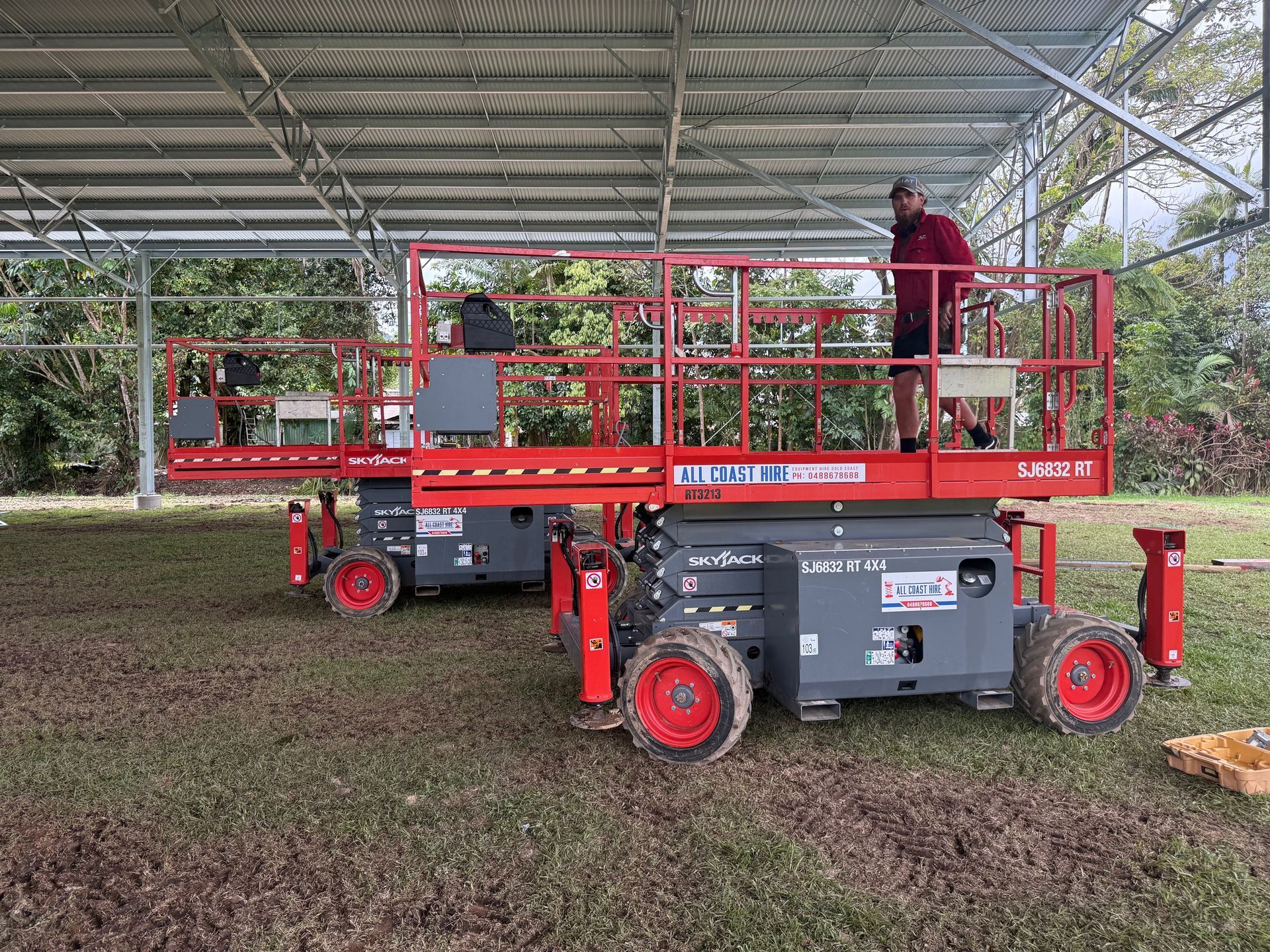 Two red and grey scissor lifts parked on a grassy field under a large metal roof structure, with a person standing on one.