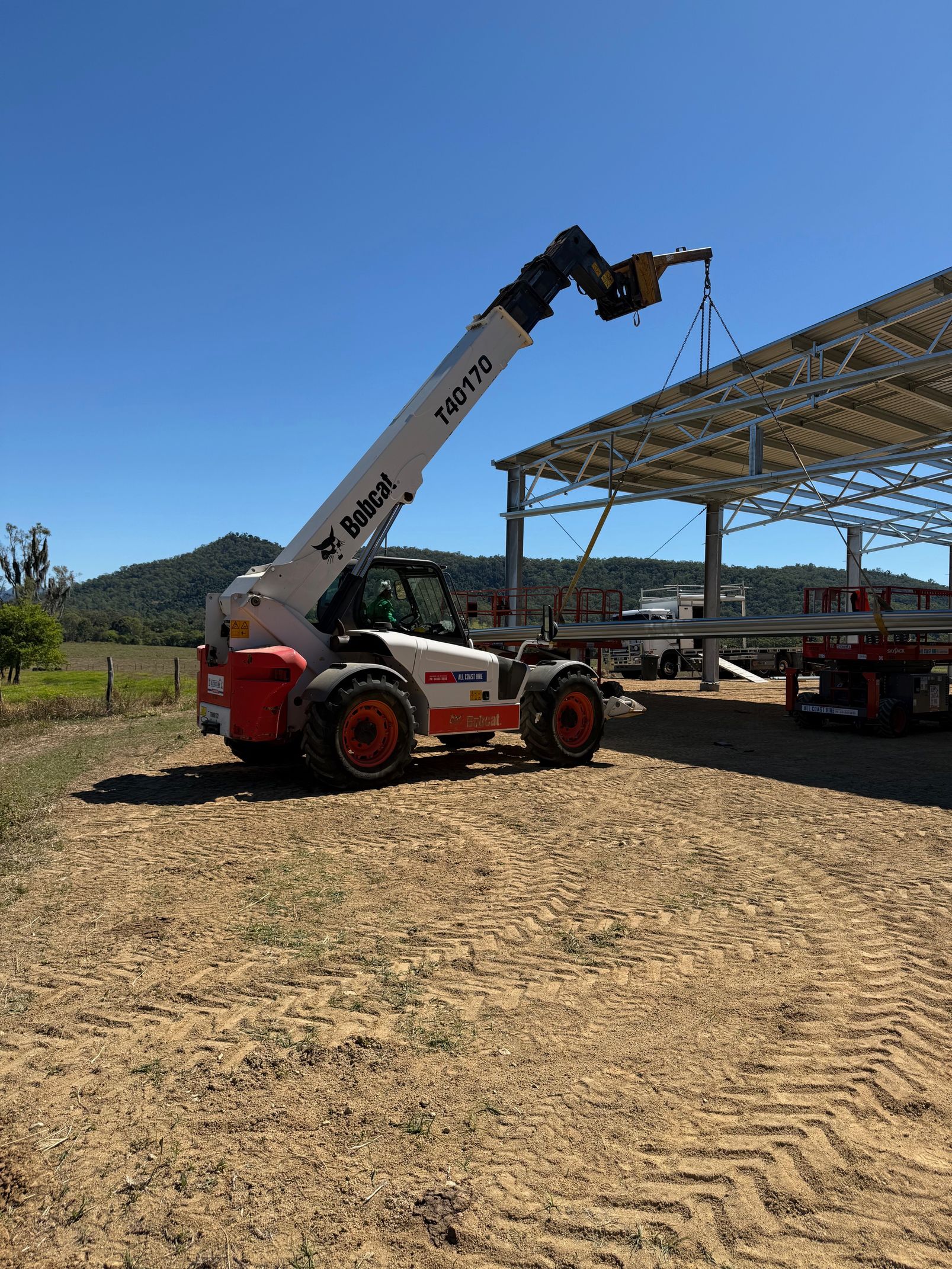 A white and orange Bobcat telehandler lifts a metal beam to construct a steel building frame under a clear blue sky.