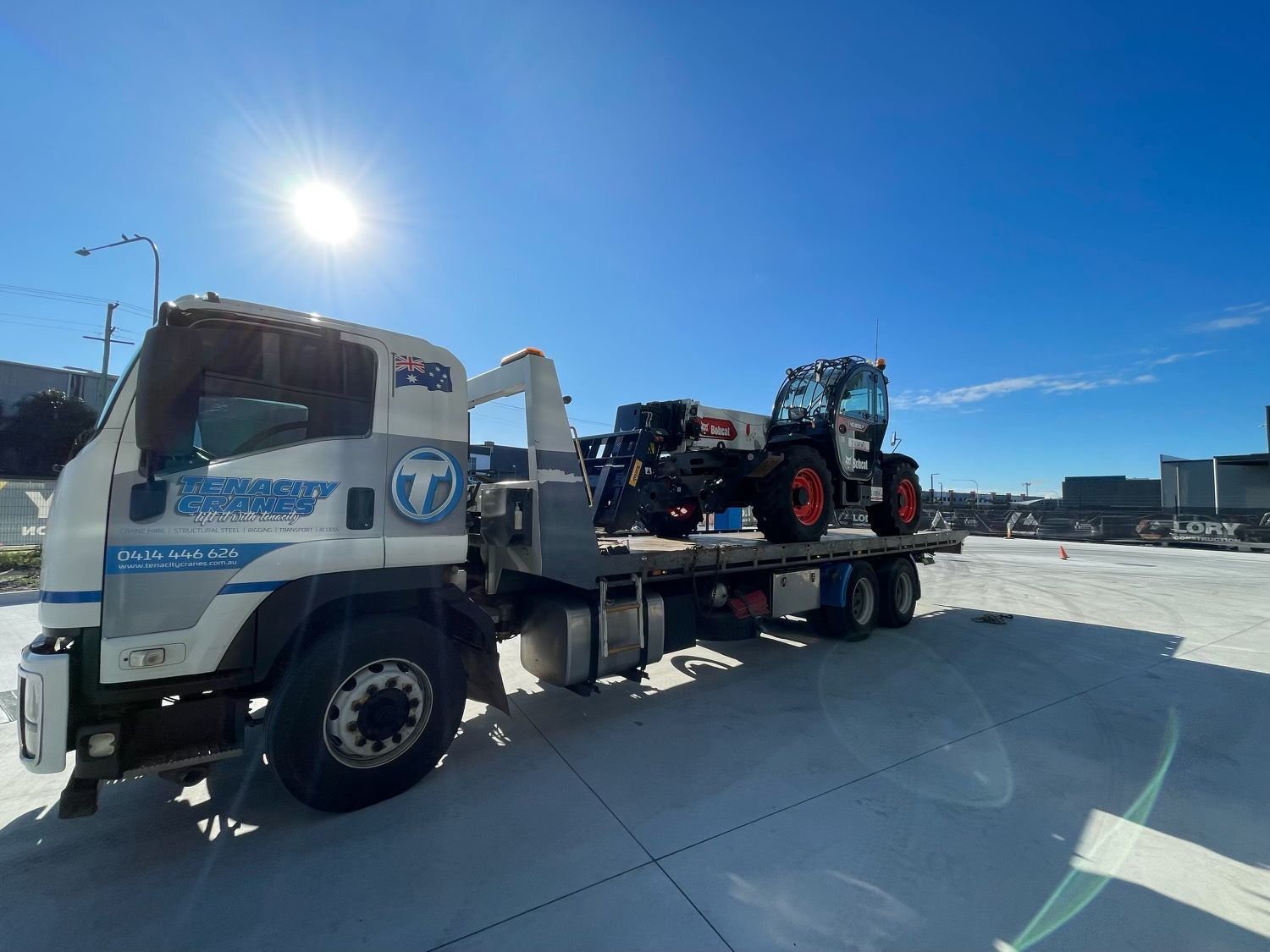 A flatbed tow truck carrying a heavy-duty telehandler vehicle on a sunny day in a paved lot.