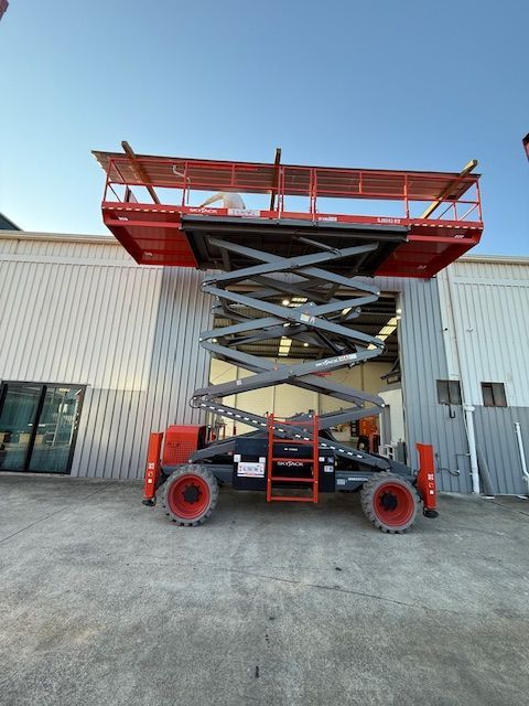 A red industrial scissor lift extended to a high position, parked on a concrete surface in front of a metal warehouse.