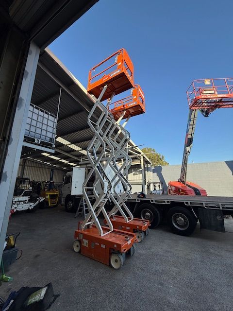 Two orange scissor lifts extended in an outdoor workshop area, one loaded on a flatbed truck.