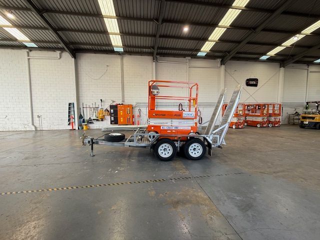 An orange scissor lift parked on a flatbed trailer inside a large, empty industrial warehouse.