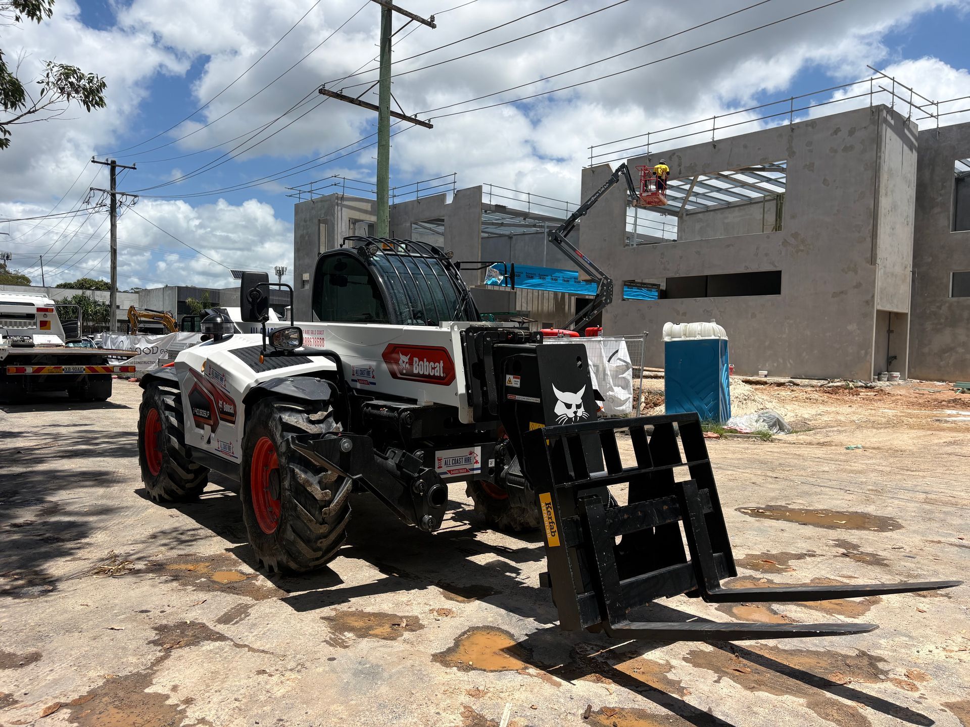 A white Bobcat telehandler with a front fork attachment parked on a dirt construction site near a concrete building shell.